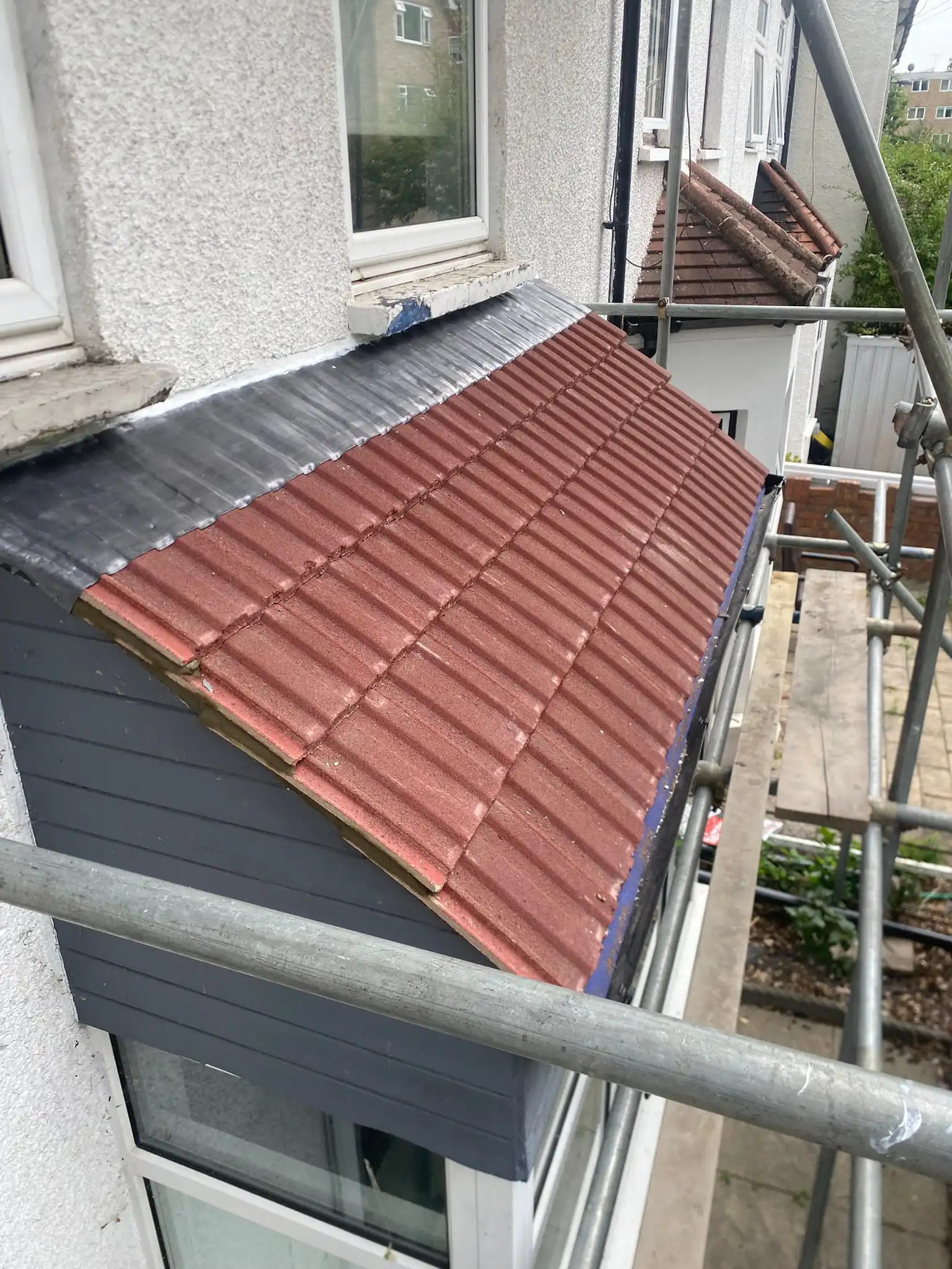 Close-up of a partially finished roof with red tiles and dark shingles, surrounded by scaffolding near a house window.