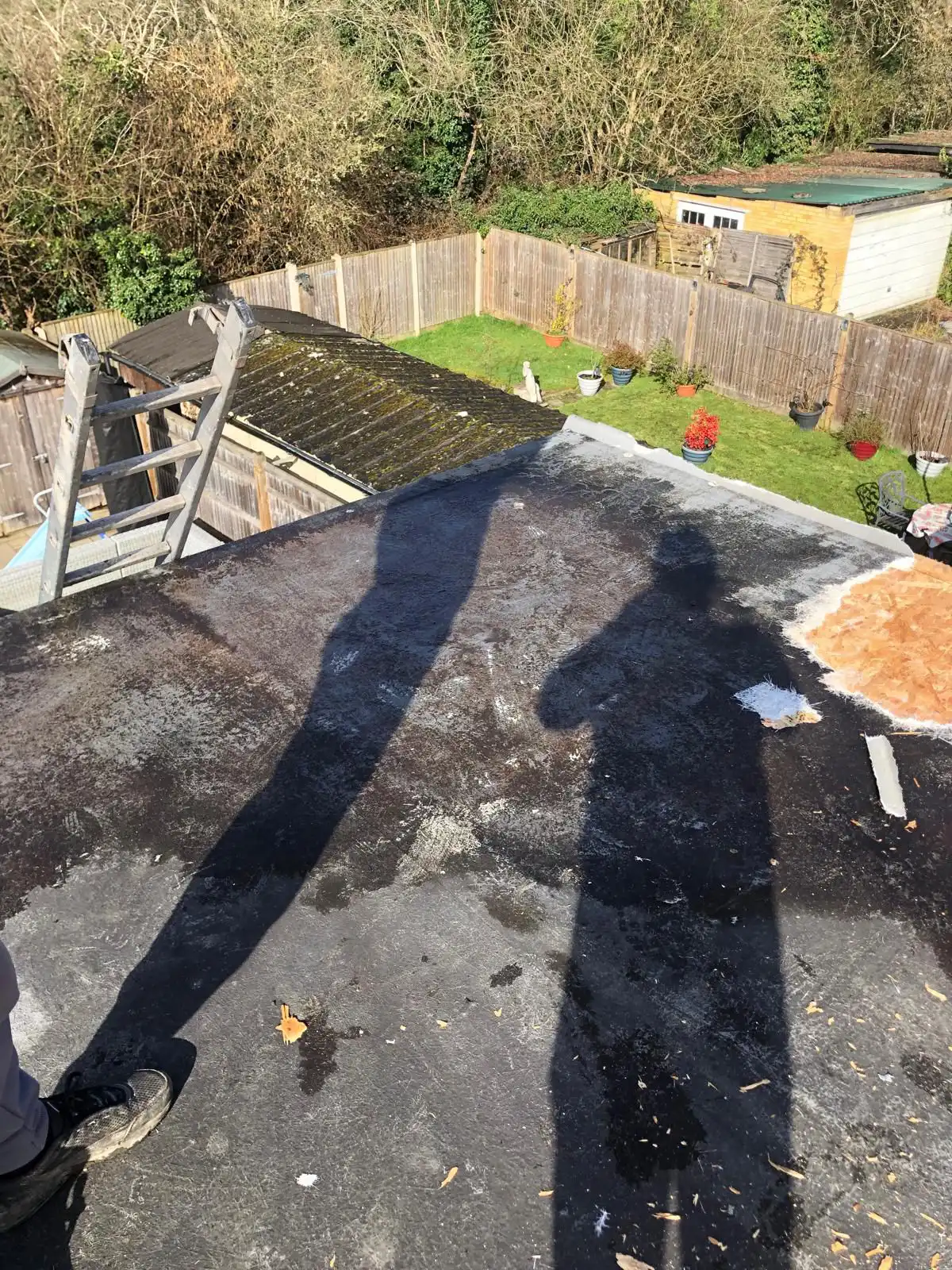 A rooftop view showing shadows on a dark surface, with a garden below featuring green grass, potted plants, and structures.
