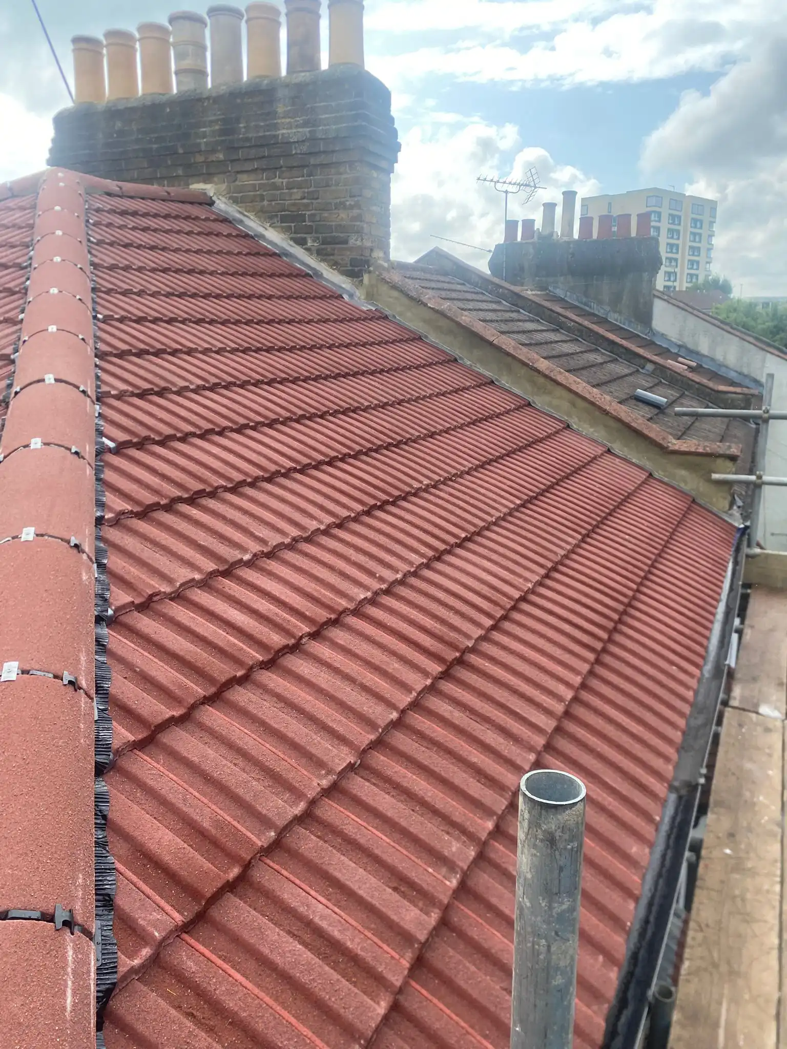 Red tiled roof with chimney stacks and a city skyline in the background. Clear skies with some clouds visible.