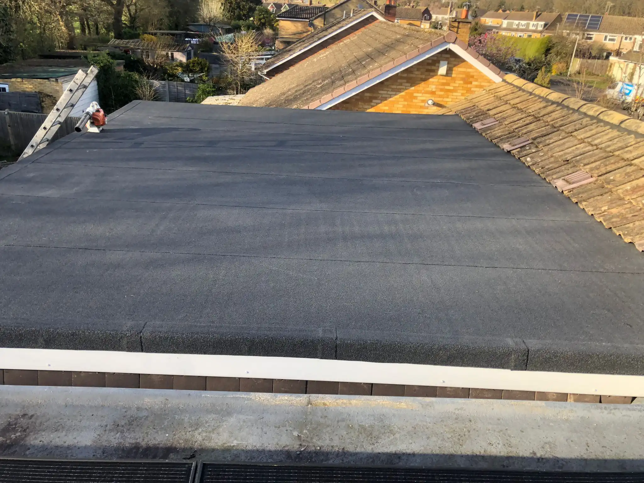Flat roof with a dark surface covering, showing a clean finish. Nearby rooftops and garden spaces are visible in the background.