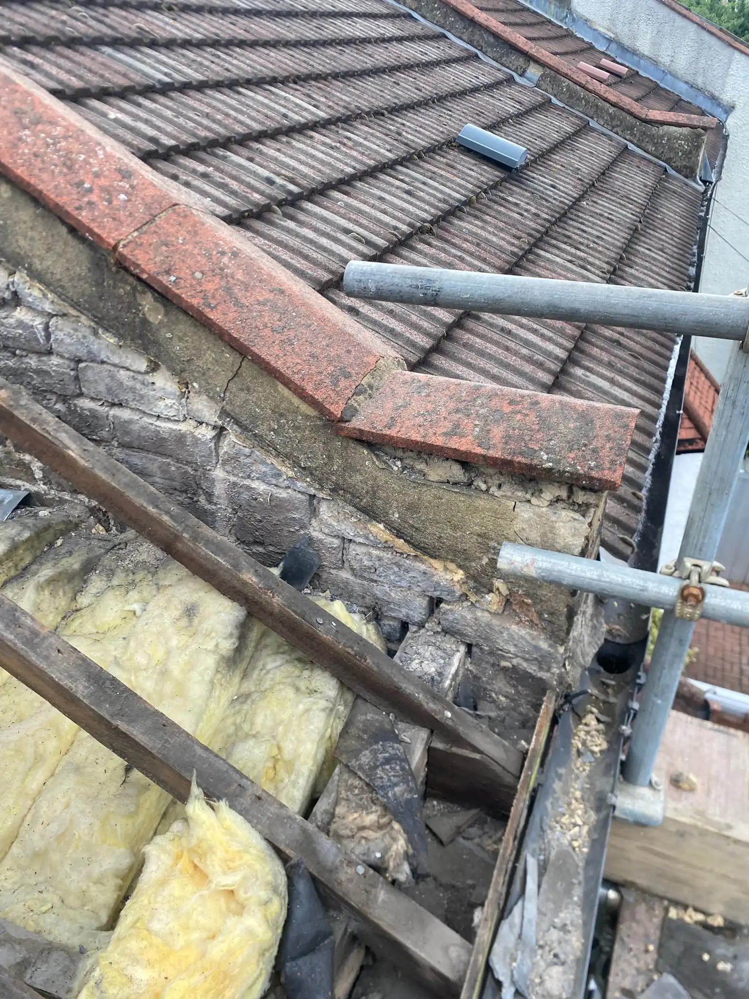 View of a roof's edge showing tiled surface, exposed insulation, wooden beams, and scaffolding for maintenance or repair.