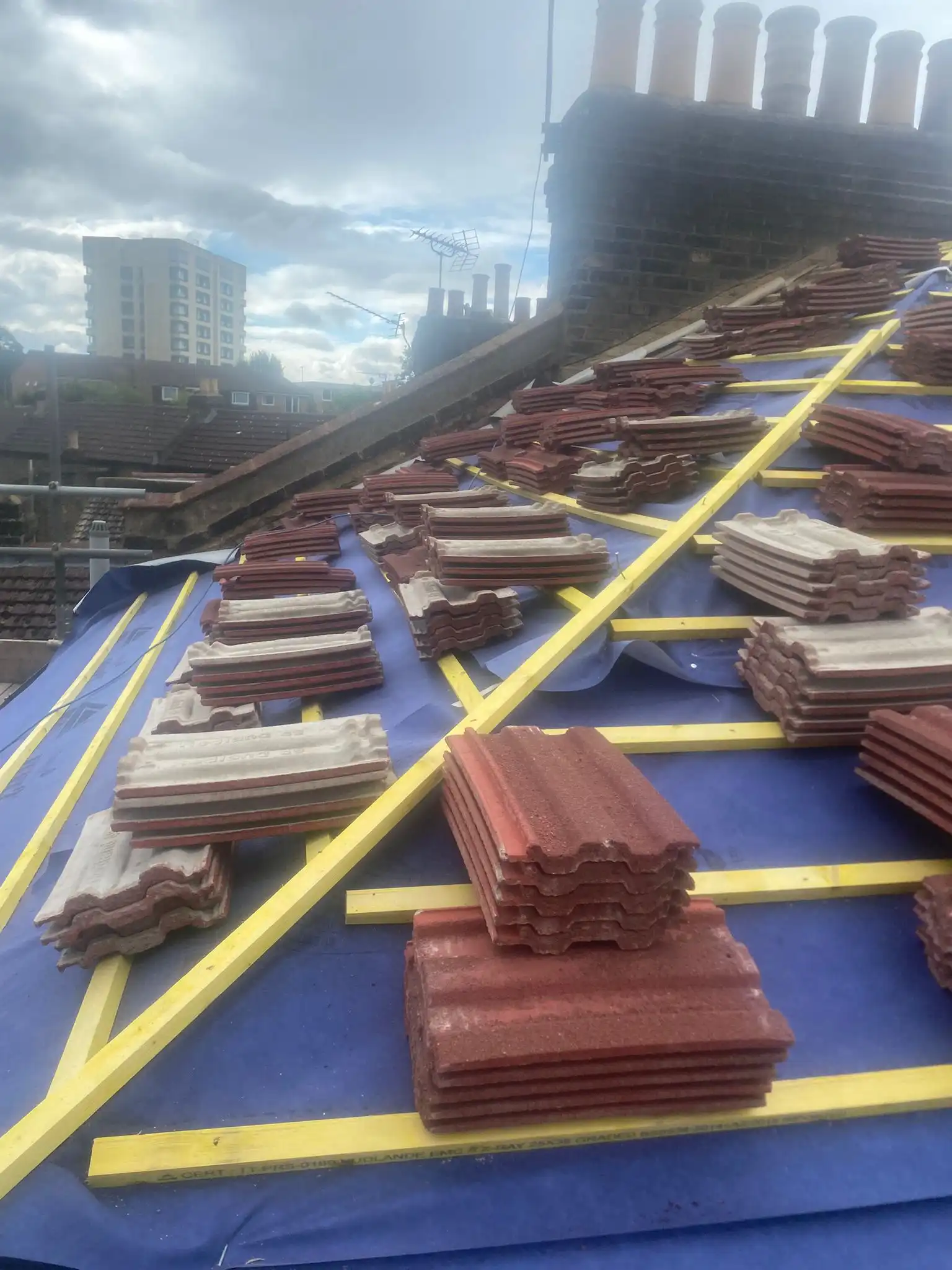 Roofing tiles stacked on a blue tarp with yellow supports, against a cloudy sky backdrop featuring a cityscape.
