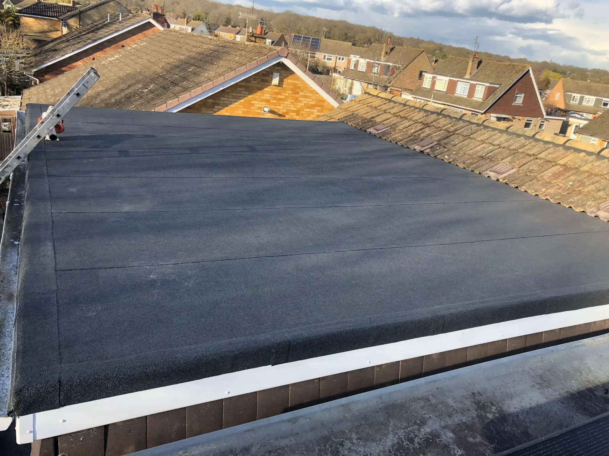Flat roof covered with black roofing material, overlooking a neighborhood with houses and trees in the background.