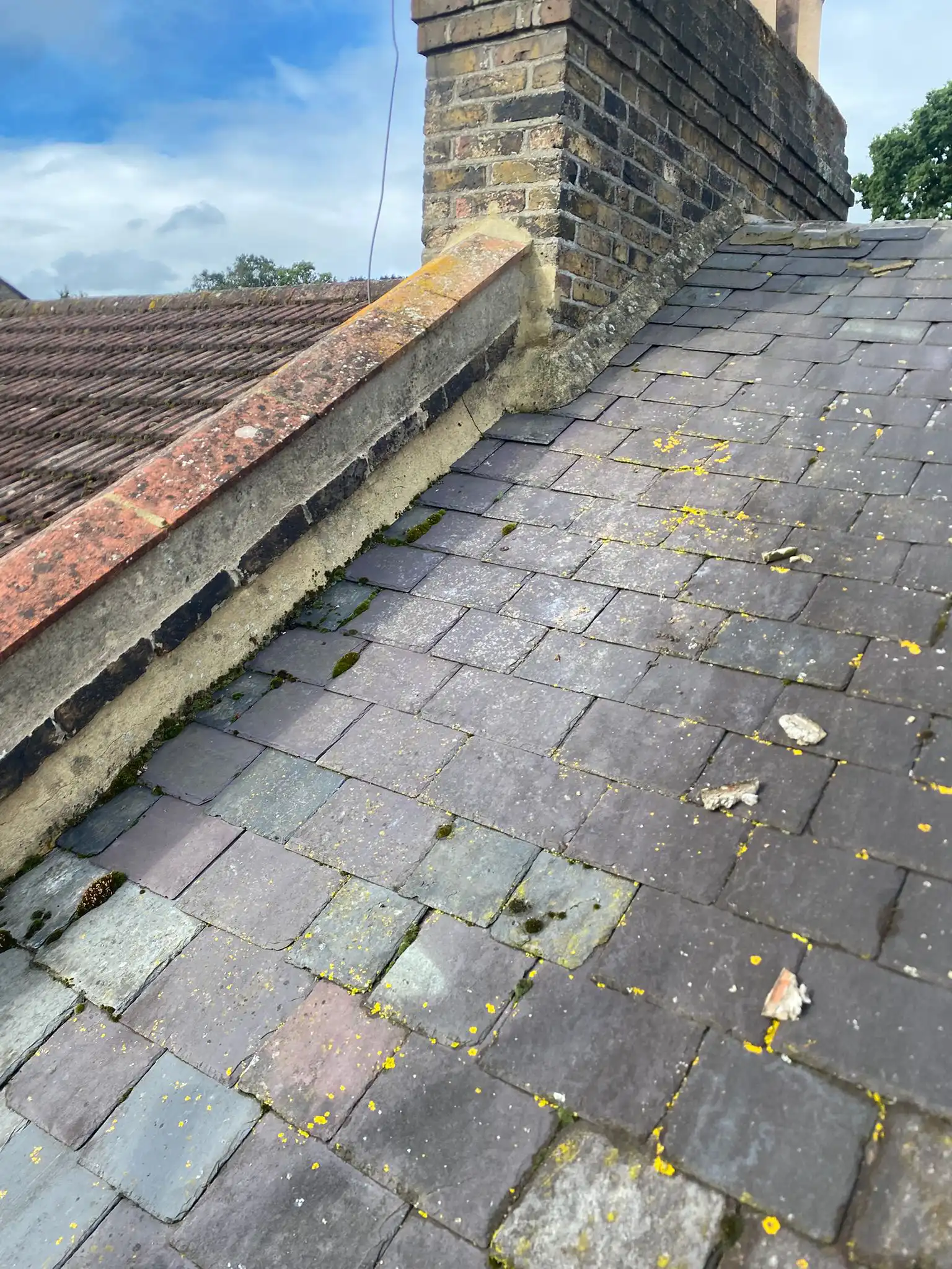 Close-up of a sloping roof with slate tiles, showing some debris, moss, and a brick chimney against a cloudy sky.