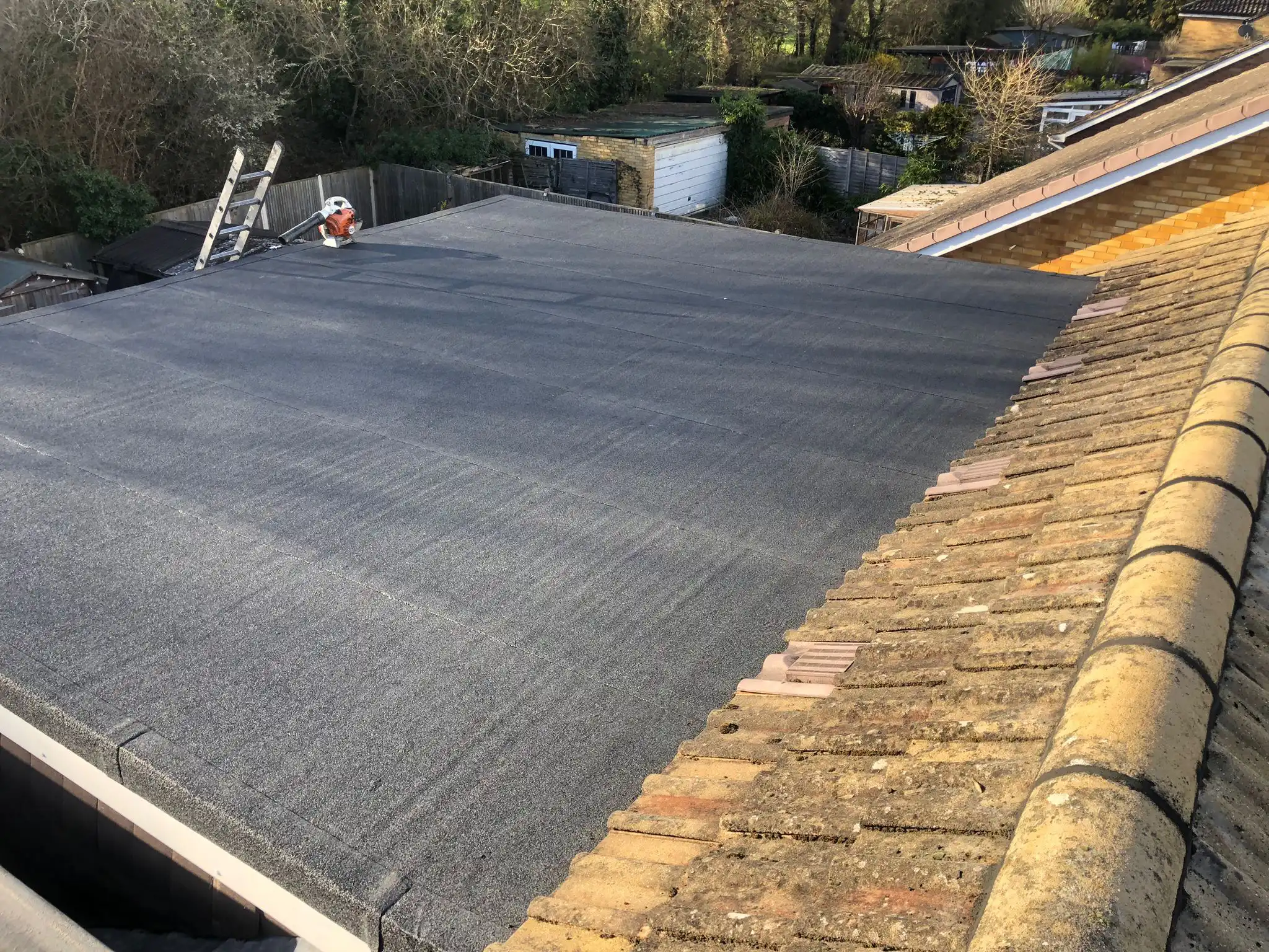 Flat roof installation in progress, featuring a black surface and a nearby ladder. Residential area with greenery in the background.