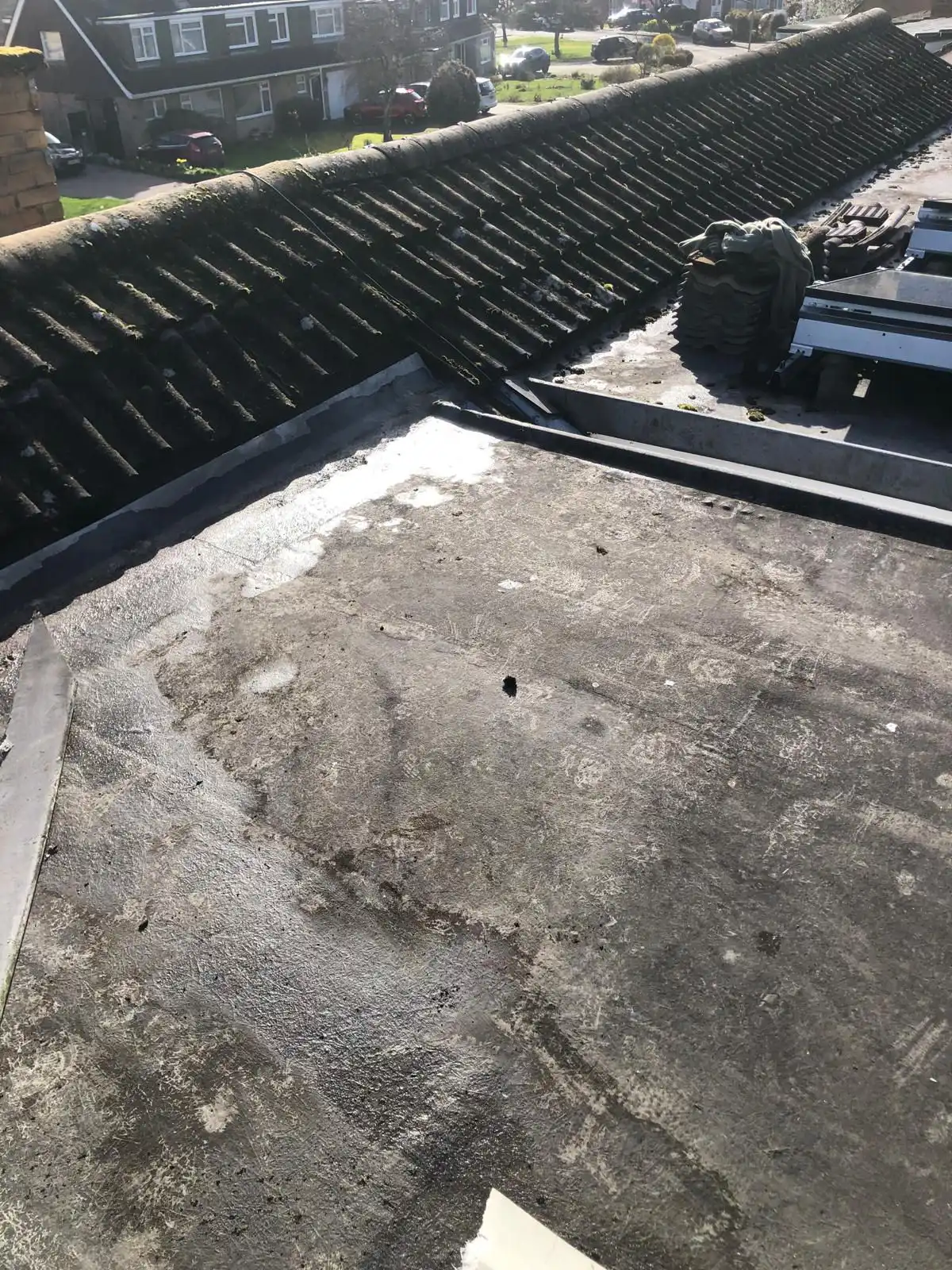 A flat roof view showing roof tiles, debris, and adjacent houses with parked cars in bright sunlight.